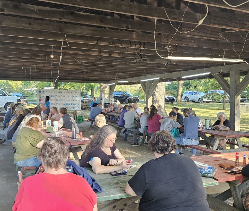 A large group of people are seated at picnic tables under a wooden pavilion in a park. They are engaged in conversation and activities, creating a lively, social atmosphere.